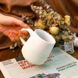 May include: White ceramic mug with a speckled glaze. The mug has a rounded shape and a handle. The mug is sitting on a magazine with text.