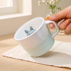 May include: A white ceramic mug with a light blue gradient at the base. A small, grey animal figure is inside the mug. The mug is held by a person.