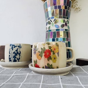 May include: Two ceramic coffee mugs with floral designs. One mug has a blue and white floral pattern, and the other has a yellow, red, and white floral pattern. The mugs are on white saucers and are sitting on a white and gray checkered tablecloth.