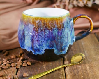 May include: A blue and brown ceramic mug with a unique glaze pattern. The mug has a rounded body and a handle. It is sitting on a wooden surface with coffee beans scattered around it.