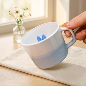 May include: A white and blue ceramic mug with a blue interior design. The mug is held by a person's hand. A small vase of daisies sits in the background.