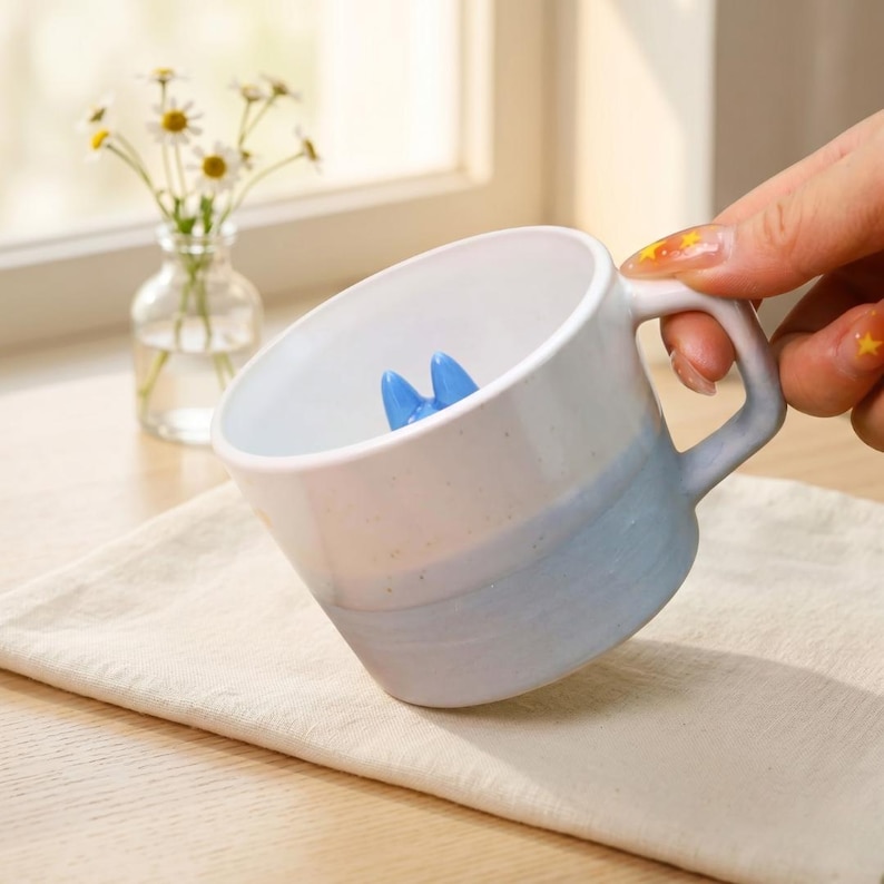 May include: A white and blue ceramic mug with a blue interior design. The mug is held by a person's hand. A small vase of daisies sits in the background.