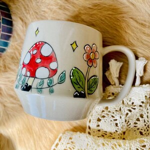 May include: A ceramic mug with a whimsical design featuring a red and white mushroom, green leaves, and a pink flower. The mug has a light gray background and a white handle.