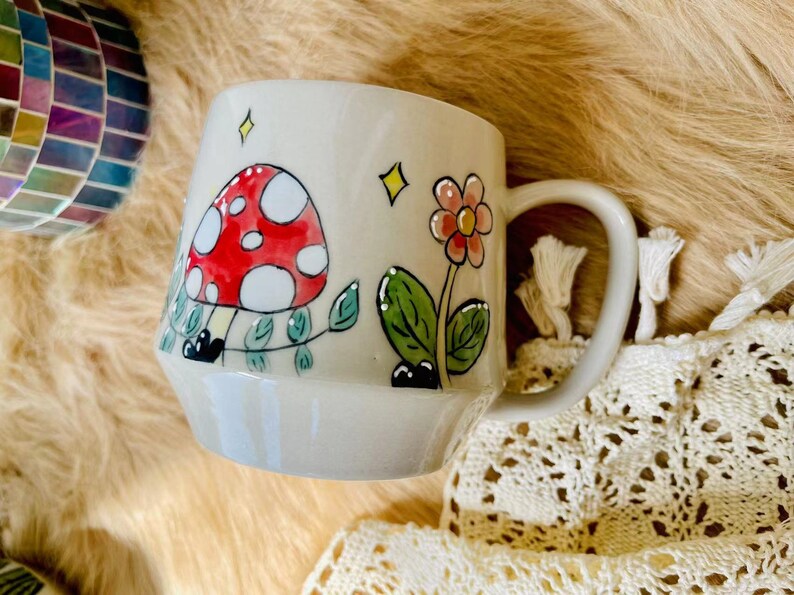 May include: A ceramic mug with a whimsical design featuring a red and white mushroom, green leaves, and a pink flower. The mug has a light gray background and a white handle.