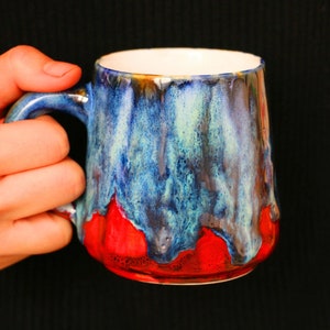May include: A blue and red ceramic mug with a unique glaze pattern. The mug has a handle and is held in a hand.