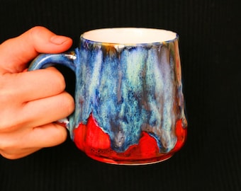 May include: A blue and red ceramic mug with a unique glaze pattern. The mug has a handle and is held in a hand.