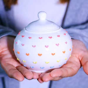 以下が含まれることがあります： カラフルなハートで飾られた蓋付きの白いセラミック製の砂糖入れ
