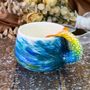 May include: A blue and green ceramic mug with a mermaid tail handle. The tail is yellow, orange, and green with scales.