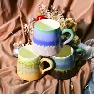 Puede incluir: Tres tazas de cerámica con diseños abstractos coloridos. Las tazas están apiladas una encima de la otra, con la taza superior de color azul claro y morado, la taza del medio de color verde y amarillo, y la taza inferior de color azul oscuro y verde.