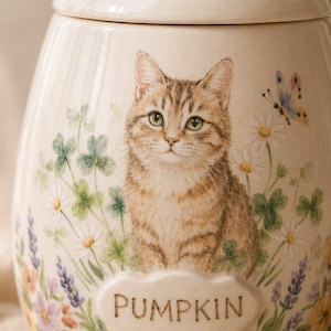 Puede incluir: Un tarro de cer&aacute;mica color crema con tapa, que presenta una ilustraci&oacute;n detallada de un gato atigrado marr&oacute;n rodeado de tr&eacute;boles verdes, margaritas blancas, lavanda morada y una mariposa. La palabra PUMPKIN est&aacute; grabada en una placa ovalada en relieve.