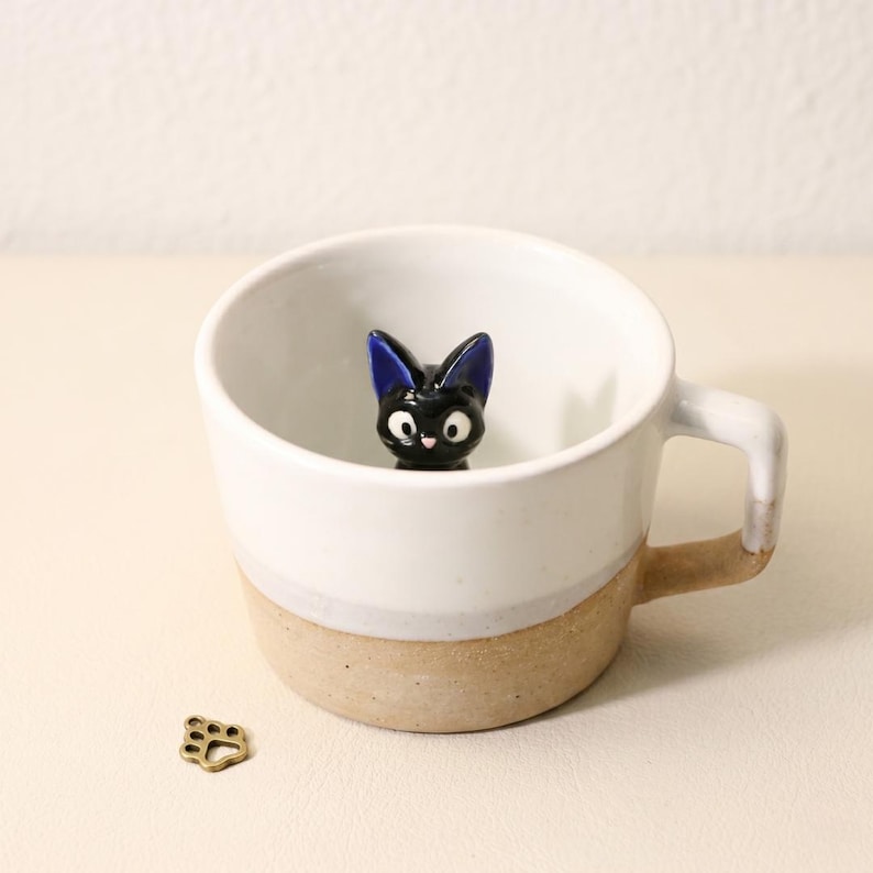May include: A white ceramic mug with a black cat figurine inside. The mug has a tan base and handle. A small, gold-coloured paw print charm is next to the mug.