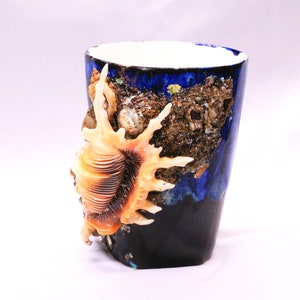Puede incluir: Una taza de cerámica azul con un diseño de concha marina. La taza tiene un interior blanco y está decorada con una concha grande en el lateral. La concha es de color marrón claro y tiene una superficie texturizada. La taza también está decorada con conchas más pequeñas y otros detalles con temática marina.