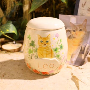 Puede incluir: Un tarro de cer&aacute;mica color crema con tapa, que presenta un gato atigrado naranja pintado a mano entre tr&eacute;boles verdes y flores peque&ntilde;as. El nombre Leo est&aacute; escrito en letras doradas sobre una placa ovalada en relieve en la parte frontal.