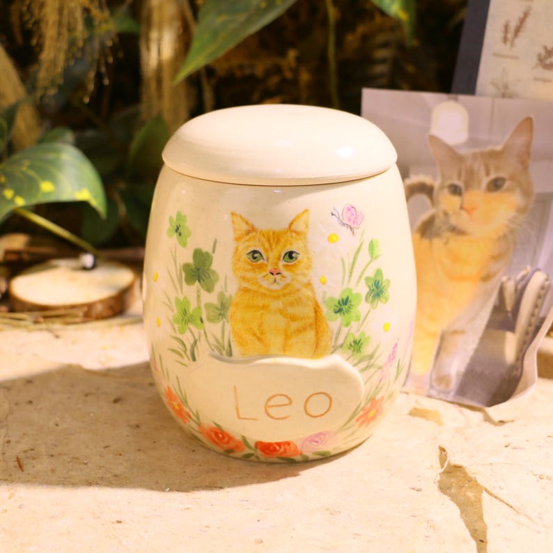Puede incluir: Un tarro de cer&aacute;mica color crema con tapa, que presenta un gato atigrado naranja pintado a mano entre tr&eacute;boles verdes y flores peque&ntilde;as. El nombre Leo est&aacute; escrito en letras doradas sobre una placa ovalada en relieve en la parte frontal.