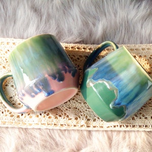 Puede incluir: Dos tazas de cerámica con un esmalte verde, azul y rosa. Las tazas tienen un aspecto único, pintado a mano y son perfectas para añadir un toque de color a tu cocina.