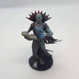 Op de afbeelding: Een blauw en grijs geverfd miniatuurbeeldje van een skeletfiguur dat een zwarte robe draagt en een open boek vasthoudt. De figuur heeft een rood en zwart stekelig kraagje en een blauw gezicht.