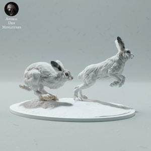 Puede incluir: Dos liebres de raqueta en miniatura pintadas de blanco, una de pie sobre sus patas traseras y la otra corriendo, sobre una base circular blanca. Animal Den Miniatures.