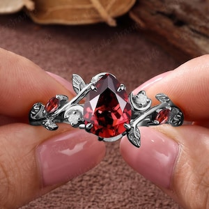 May include: A black metal ring featuring a central pear-cut deep red gemstone, accented by smaller red stones and clear white stones set within a leaf-shaped band design. The ring has a dark finish and intricate botanical-inspired metalwork.