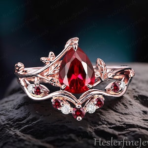Puede incluir: Anillo de oro rosa con una piedra preciosa roja oscura en forma de pera. El anillo presenta detalles de hojas y vides, y piedras preciosas rojas y blancas más pequeñas. El anillo está sobre una superficie oscura y texturizada.