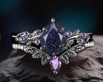 May include: A pear-shaped blue gemstone ring with a black metal band and delicate leaf-like details. The ring is set with smaller purple gemstones and has a matching band with white gemstones.