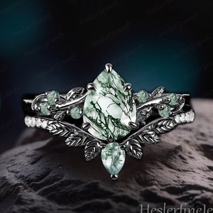 May include: A black and green engagement ring set with a pear-shaped green moss agate gemstone. The ring features a delicate leaf design with smaller green moss agate accents.