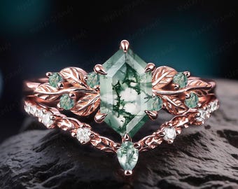 May include: A rose gold ring set featuring a large, faceted green gemstone with a unique pattern. The set includes a leaf-adorned band and a diamond-studded band, creating an elegant and nature-inspired design. Heslerfinejewelry is printed on the bottom right.