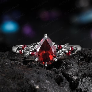Könnte beinhalten: Ein Ring mit einem birnenförmigen, dunkelroten Edelstein in einer silberfarbenen Metallschiene. Die Schiene zeigt ein rankenartiges Design mit kleinen Blättern und mehreren winzigen, runden roten Akzentsteinen, die seitlich eingearbeitet sind.