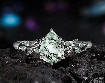 May include: A silver-toned ring featuring a pear-shaped moss agate center stone with dark green inclusions. The band is designed with metal leaf motifs and small round moss agate accents on both sides of the central pear-shaped gemstone.