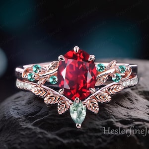 May include: A rose gold engagement ring set with a large oval red gemstone and smaller green gemstones set in a leaf design. The ring is on a grey rock.