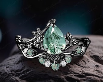 Könnte beinhalten: Ein schwarzer Ring mit einem birnenförmigen grünen Edelstein, der von kleineren grünen und weißen Edelsteinen umgeben ist. Der Ringband ist so gestaltet, dass er wie eine Rebe mit Blättern aussieht.