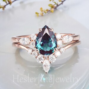 May include: A pear-shaped green and blue gemstone ring with a halo of smaller white gemstones. The ring is set in rose gold and has a matching band with a curved design.