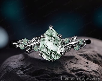 May include: A pear-shaped green moss agate engagement ring with a silver band and small green stones on each side. The ring is set on a dark gray rock.