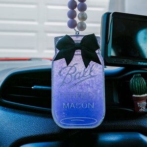 May include: Purple glass jar car air freshener with a black bow and the text "Ball Perfect Mason" hanging from a rearview mirror with a string of purple and white beads.