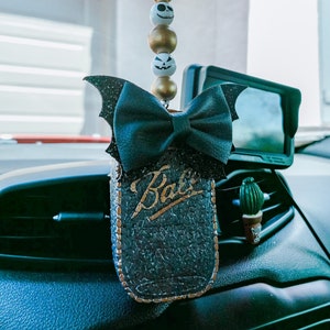 May include: A black and gold car air freshener with a black bow and bat wings. The air freshener is shaped like a mason jar with the word "Bali" written on it. The air freshener is hanging from the rearview mirror of a car.