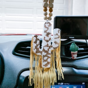 Puede incluir: Un ambientador de coche en forma de cactus blanco y dorado con una borla dorada que cuelga de la parte inferior. El cactus está decorado con un estampado de leopardo. El ambientador está colgado de un espejo retrovisor en un coche.
