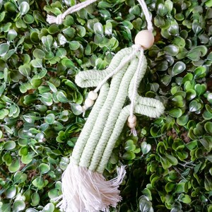 Op de afbeelding: Een lichtgroen macramé cactusornament met een witte kwast en houten kralen. De cactus heeft een ophanglus en staat tegen een achtergrond van groen gebladerte. Dit decoratieve item is geschikt voor woondecoratie.