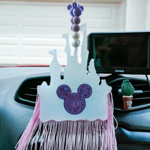 Puede incluir: Un ambientador de coche blanco con forma de castillo de Cenicienta con una cabeza de Mickey Mouse de purpurina morada en el centro. El ambientador tiene flecos rosas alrededor de la parte inferior y está colgado de un espejo retrovisor.