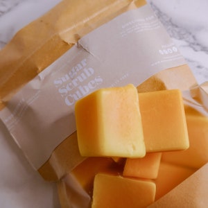 May include: A brown paper bag with a white label that says "Sugar Scrub Cubes" and contains several bright orange sugar scrub cubes.