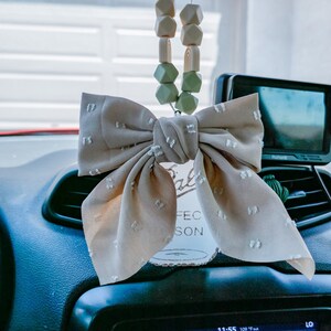 May include: A beige fabric bow with white polka dots hangs from a rearview mirror in a car. The bow is attached to a string of green and beige silicone beads. The car's dashboard is visible in the background.