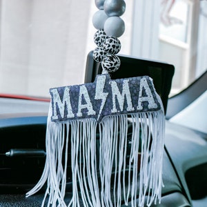 May include: Car hanging ornament featuring the word "MAMA" in a decorative font, with a lightning bolt design. The ornament has white fringe and a string of decorative beads in silver, grey, and cow print.