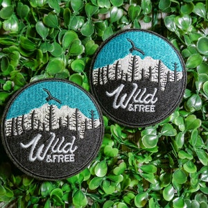 Op de afbeelding: Twee gestikte zwarte en turquoise patches met een berglandschap en de tekst "Wild & Free".