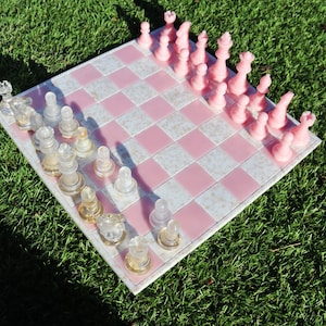 May include: A pink and white chessboard with pink chess pieces on one side and clear glass chess pieces on the other side. The chessboard is sitting on green grass.