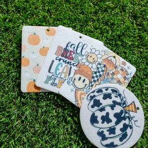 Pode incluir: Três aromatizadores de carro brancos com designs de outono. Um apresenta uma abóbora com um padrão de vaca, outro tem um rosto sorridente com o texto "fall breeze crunch leaves pumpkin pie", e o último tem um padrão de abóboras laranja sobre um fundo branco.