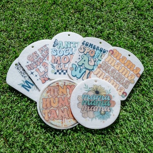 Könnte beinhalten: Sechs verschiedene Auto-Lufterfrischer mit verschiedenen Designs und Texten. Die Designs umfassen ein Blumenmuster, ein Regenbogenmuster und ein kariertes Muster. Der Text umfasst "On My Husband", "Anti Social Mom Club", "Somebody Spoiled Me", "Mama Mama Mama", "Tiny Human Family" und "Mama Mama Mama".