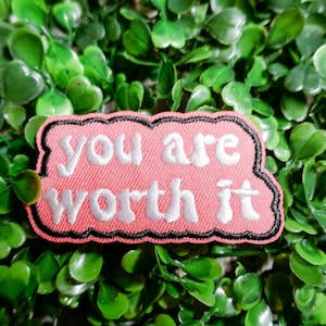 Può includere: Un cerotto in tessuto rosa con un contorno nero e un testo ricamato bianco che dice "you are worth it".