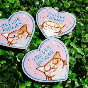 May include: Three pink heart-shaped embroidered patches with a white cat and the text "You are enough" in white. The patches have a blue outline and are decorated with small white diamonds.