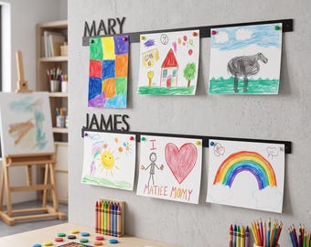 以下が含まれることがあります： MARYとJAMESという名前が切り抜かれた、黒い金属製の水平な壁掛けディスプレイレール2本。各レールには小さなカラフルなマグネットで子供の絵が3枚ずつ留められており、クレヨンで描かれた様々なイラストが飾られています。