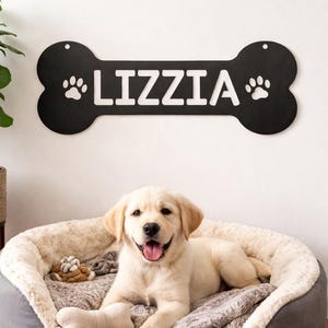 May include: A smiling golden Labrador puppy rests in a plush, round dog bed with a gray exterior and a cream-colored, fluffy interior. Above, a black bone-shaped sign reads "LIZZIA" with paw prints.