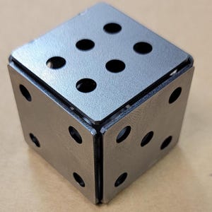 May include: A metal dice with six sides, each side has a different number of holes. The dice is made of a silver-colored metal and has a textured surface.