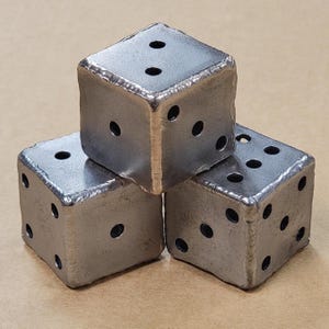 May include: Three metal dice, each with six sides and holes for each number. The dice are made of a dark gray metal and are arranged in a triangular shape.